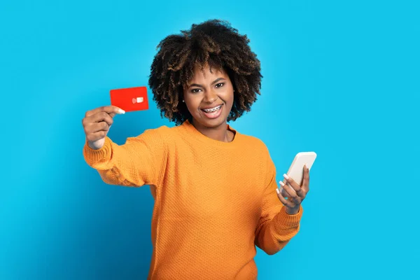 快乐的微笑迷人的年轻黑人女性 留着浓密的头发和牙齿 手里拿着红卡和手机 在网上购买蓝色的工作室背景 复制空间 — 图库照片