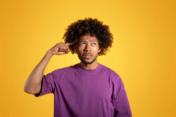 Pensive sad confident adult african american curly man in purple t-shirt puts his finger to temple isolated on yellow studio background. Brainstorm, creative idea, think and choice