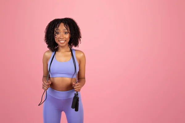 スポーツウェアを着た若いアフリカ系アメリカ人女性の笑顔がジャンプロープを保持し ピンクのスタジオの背景だけでスポーツをお楽しみください アクティブなライフスタイル 心臓トレーニング ボディケアとワークアウトをフィット — ストック写真