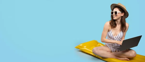 Young lady in swimsuit, straw hat and sunglasses sitting on inflatable mattress and using laptop, looking aside at copy space on blue background, panorama