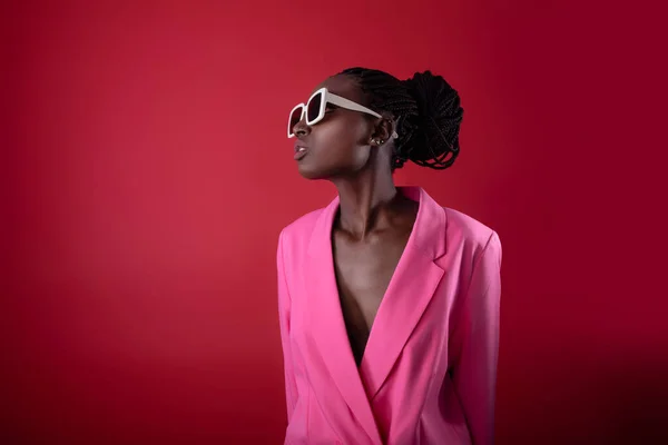 Beautiful African American Woman In Stylish Fashionable Clothes Posing On Red Background, Young Black Woman Wearing Pink Blazer Jacket And Stylish Eyeglasses Looking Aside At Copy Space