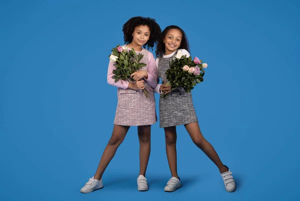 stock image Cheerful cute black little girls girlfriends with bouquets of flowers, enjoy greeting birthday, isolated on blue background, studio. Holiday celebration, congratulation, childhood and emotions
