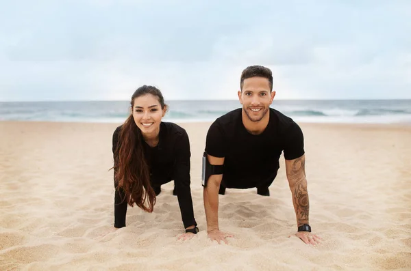 Happy fit couple in sportswear do push ups or plank on ocean beach, exercising outdoors and smiling at camera. Relationship, health care, active lifestyle concept