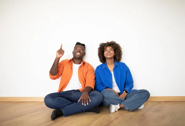 Great Idea. Happy Black Couple Sitting On Floor Near Wall In New Flat And Pointing Up, Smiling Young African American Spouses Thinking About Decorations And Furniture After Moving Home, Copy Space