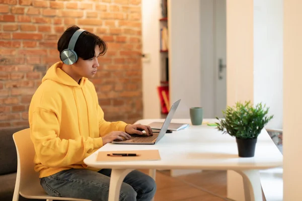 オンライン学習 ラップトップを使用して中国の学生の男宿題をし 自宅でヘッドフォンを着てインターネットサーフィン 10代の男の子が机の上に座ってコンピュータを入力してのサイドビューショット屋内 — ストック写真