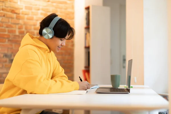Eラーニング ノートパソコンを使ったアジア系学生のサイドビューとメモを取るライティング 自宅でオンラインクラスを持っている 机の上に座っているワイヤレスヘッドフォンを身に着けている幸せな十代の若者 — ストック写真