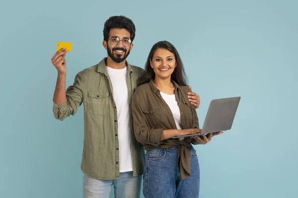 黄色の銀行カードを示すカジュアルな衣装で陽気な幸せな正の若いインドのカップルと青のスタジオの背景にラップトップを使用して 恋人のショッピングやインターネット上の銀行 オンライン決済 — ストック写真