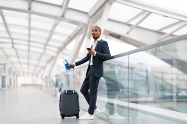 Uçuşu bekliyorum. Havalimanında akıllı telefon kullanan genç siyahi işadamı, Afrika kökenli, takım elbiseli, pasaportlu ve terminalde dururken bavulunu taşıyan Amerikalı bir erkek.