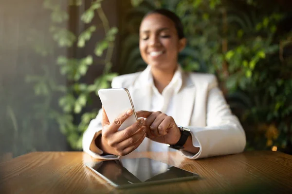 Glad young black woman in suit chatting on phone at table with tablet with empty screen in cafe with plants interior. Website, blog, video call modern app for business and work