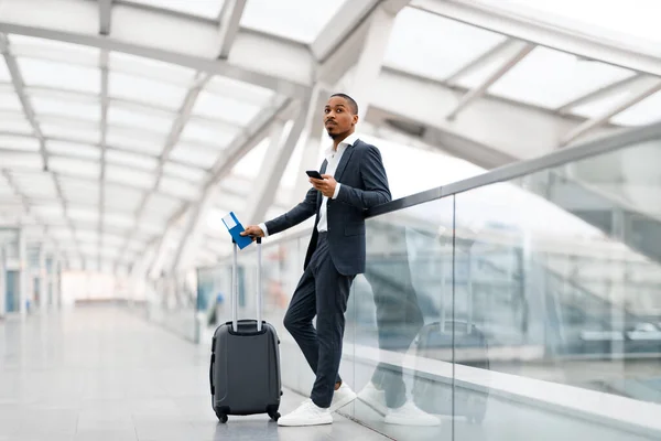 飛行を待っている 空港でスマートフォンを使用している若い黒人ビジネスマン チケットでスーツケースを保持し ターミナルに立ちながらスーツケースを運ぶでハンサムなアフリカ系アメリカ人男性 — ストック写真