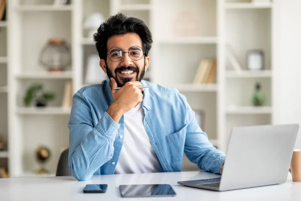 Successful Freelancer. Happy Handsome Indian Man Posing At Desk With Laptop, Young Eastern Male Touching Chin And Smiling At Camera While Working With Computer At Home Office, Copy Space