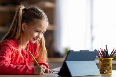 Online Eğitim Konsepti. Evde dijital tablet kullanan sevimli küçük bir kızın portresi, uzaktan çalışma için modern aletleri kullanan gülümseyen liseli kız çocuğu, deftere notlar yazma, yer kopyalama.