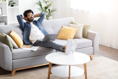 Hayalperest genç Hintli adam ev içinde dizüstü bilgisayarla rahatlıyor, yakışıklı, serbest çalışan, elleri başının arkasında koltuğa yaslanan, başarılı bir çevrimiçi çalışmadan sonra dinleniyor, boşluğu kopyalıyor.