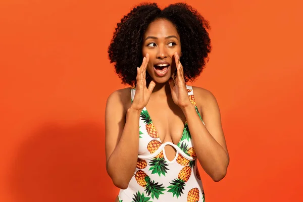 stock image Travelling deal. Positive attractive young curly black woman in colorful swimsuit holding hands next to mouth and looking aside, making announcement on orange studio background, copy space