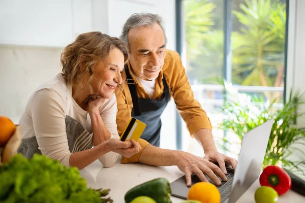 台所の内部でラップトップとクレジットカードを使用して興奮した高齢者の配偶者 高齢者のカップルは オンライン食料品や食品の配達を注文します 自宅でコンピュータ経由でインターネットショッピング — ストック写真