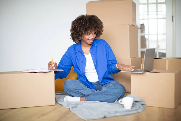 Young black woman taking notes and using laptop while in her new house after moving, smiling african american female sitting among cardboard boxes with belongings, making checklist for relocation