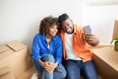 Mutlu Afrika kökenli Amerikalı çift eve taşınırken akıllı telefondan selfie çekiyor, neşeli siyah eşler yeni apartman dairelerinde karton kutuların arasında oturuyor ve fotoğraf makinesine poz veriyor, taşınmayı kutluyor.