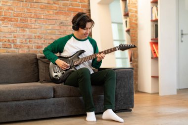 Mutlu Çinli genç adam müzik öğreniyor. Elektro gitar çalıyor ve kulaklık takıyor. Yetenekli müzisyen çocuk evde koltukta oturan müzik aleti ile poz veriyor. Hobi ve Boş Zaman