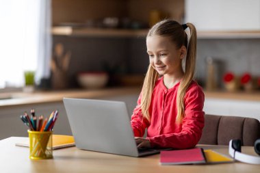 Evde eğitim kavramı. Evde dizüstü bilgisayar kullanan tatlı bir kız, masada oturan ve bilgisayar başında çalışan bir kız, cihaz ekranına bakıyor, uzaktan öğrenmenin keyfini çıkarıyor, boşluğu kopyalıyor.