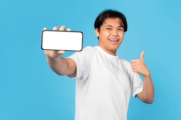 青いスタジオの背景の上に空のスクリーンジェスチャー親指でスマートフォンを示す陽気なアジアのティーンの男の子 カメラに微笑む 男の広告と承認モバイルオファー モックアップ — ストック写真