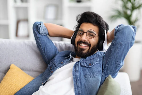 英俊潇洒的印度小伙子 戴着无线耳机 在家里听音乐 戴着眼镜的年轻快乐的东方人 在客厅里沙发上放松 喜欢最受欢迎的演奏家 闭门造车 — 图库照片
