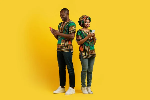 幸せな黒恋人中年男性と若い女性で全国的なアフリカの衣装でスマートフォンを使用して黄色のスタジオの背景に チャット 使用して日付アプリ フル長さ コピースペース — ストック写真
