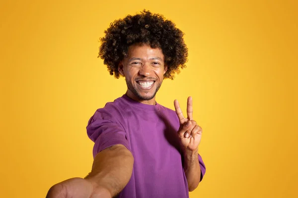 身穿紫罗兰色T恤的积极的非洲裔美国卷曲男子手拿着和平标志 带着黄色工作室背景的自拍 照片为社交网络 伟大的新闻情绪 广告和提供 — 图库照片