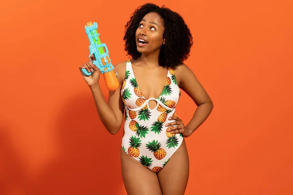 穿着时髦的游泳衣 拿着水枪 抬起头 在橙色工作室的背景上玩乐 模仿空间的漂亮而又酷的年轻黑人女人 暑假期间的水活动 — 图库照片