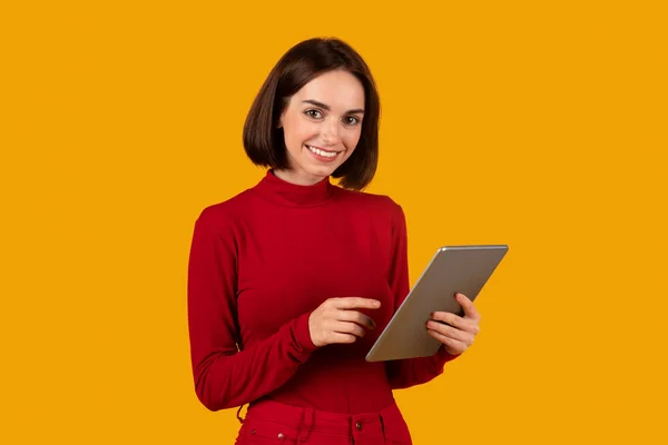 陽気です可愛いですブルネット若い女性で赤デジタルノマドで現代的なPcデジタルタブレットを使用してオレンジスタジオの背景に タッチパッドの画面と笑顔 楽しい素敵なアプリケーション コピースペース — ストック写真