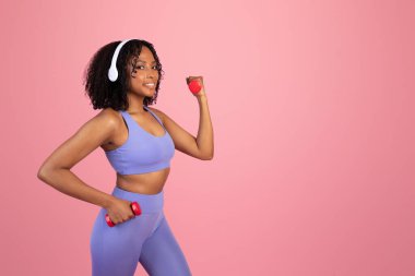Spor kıyafetleri ve kablosuz kulaklıklar takan, halterlerle el egzersizi yapan, pembe stüdyo arka planında müzikle eğlenen mutlu siyah kadın. Formda yaşam tarzı, vücut bakımı ve egzersiz