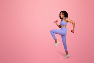 Spor giyimli neşeli, Afro-Amerikan bayan. Kol halterleriyle kardiyo egzersizi yapıyor. Pembe stüdyo arka planında tek başına sporun tadını çıkarıyor. Formda yaşam tarzı, reklam ve teklif, vücut bakımı
