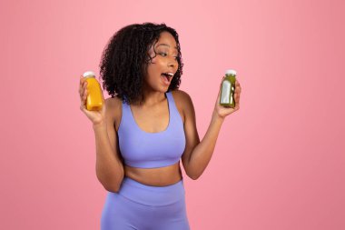Mutlu gülen Afro-Amerikan kıvırcık kadın spor giyim kuşamında ya da meyve suyu şişelerinde, pembe stüdyo arka planında izole edilmiş. Formda, aktif yaşam tarzı, vücut bakımı, diyet ve sağlıklı içecek.