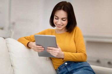 Mutlu, gevşemiş, esmer, güzel saç stili olan, rahat giyimli, kanepede oturan, dijital tablet kullanan, internette sörf yapan, e-kitap okuyan, uzay kopyası yapan güzel bir kadın.