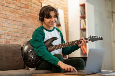 Mutlu Koreli adam elinde elektro gitar tutuyor dizüstü bilgisayarında düğmeye basıyor, evde şarkı çalıyor ve kaydediyor. İnternette Müzik Enstrümanı Çalmayı Öğrenen Genç Müzisyen Akortlarını Arıyor