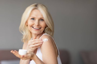 Güzel, olgun bir bayan vücut losyonu kullanıyor ya da duş aldıktan sonra omzuna nemlendirici sürüyor ya da krem sürüyor, kameraya bakıyor ve gülümsüyor, boşluğu kopyalıyor.