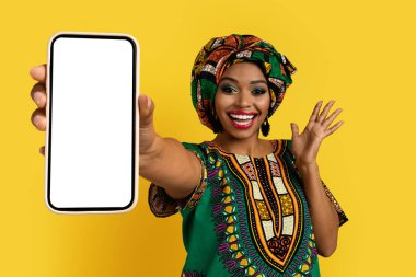 Harika bir online teklif. Mutlu duygusal gülümseme, parlak Afrika kostümü içindeki çekici siyahi bayan cep telefonunu gösteriyor ve sarı stüdyo arka planında hareketler yapıyor.
