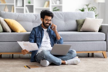 Hintli serbest çalışan adam dizüstü bilgisayar kullanıyor ve cep telefonuyla konuşuyor, gözlüklü Doğulu adam oturma odasında otururken uzaktan bilgisayar ve kağıtlarla çalışıyor, uzay kopyaları yapıyor.