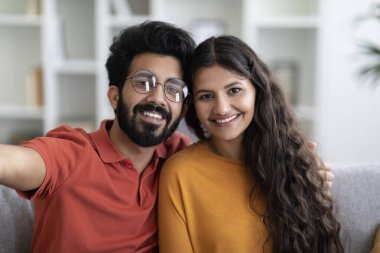 Mutlu genç Hintli çiftin evde selfie çekerken çekilmiş yakın plan portresi, mutlu yüzyıllık doğu erkek ve kadın kucaklaşıp gülümsüyor, oturma odasında poz veriyorlar.