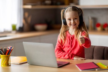 Evde dizüstü bilgisayarı olan ve kamerada baş parmağını gösteren, bilgisayar ve çalışma kitaplarıyla masada oturan, kablosuz kulaklık takan, neşeli, küçük bir kız çocuğu.