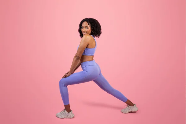 脚のストレッチ運動を行うスポーツウェアで若いアフリカ系アメリカ人女性の笑顔 ピンクのスタジオの背景だけでスポーツを楽しむ フル長さ フィットライフスタイル ボディケアとワークアウト — ストック写真