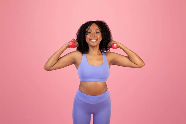 若いアフリカ系アメリカ人女性がダンベルと手のための運動をするスポーツウェアで ピンクのスタジオの背景だけでスポーツをお楽しみください アクティブなライフスタイル ボディケア ジムでのワークアウトをフィット — ストック写真