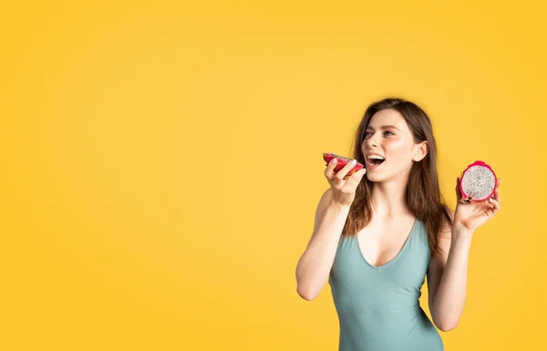 身穿泳衣的年轻貌美的女士咬着豆蔻片 摆设着龙果的姿势 享受着美味多汁的水果 站在黄色的工作室背景上 享受着自由的空间 — 图库照片