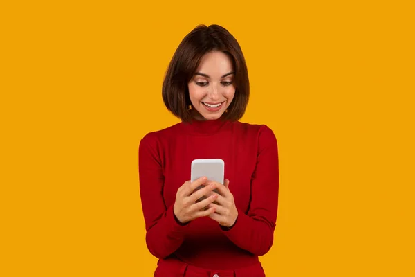 穿着红色休闲装 情绪愉悦 美丽的黑发女青年 在橙色工作室的背景下 用白色的现代手机 看着智能手机屏幕 微笑着 享受着伟大的移动应用 — 图库照片