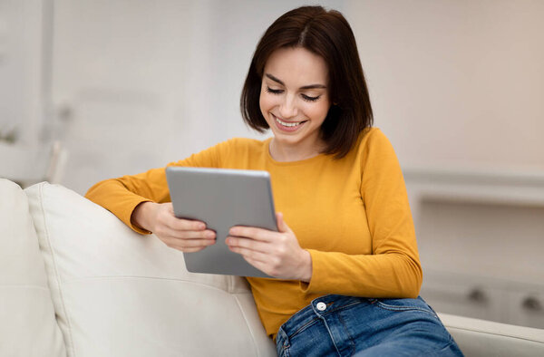 Happy relaxed beuatiful brunette young woman with nice hairstyle wearing casual outfit sitting on couch, using digital tablet at home, surfing on Internet, reading e-book, copy space