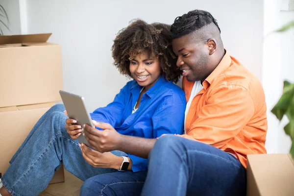 Happy Black Couple Using Interior Design App On Digital Tablet After Moving Home, Smiling African American Spouses With Modern Gadget Planning Decorations In New Apartment, Sitting Among Boxes