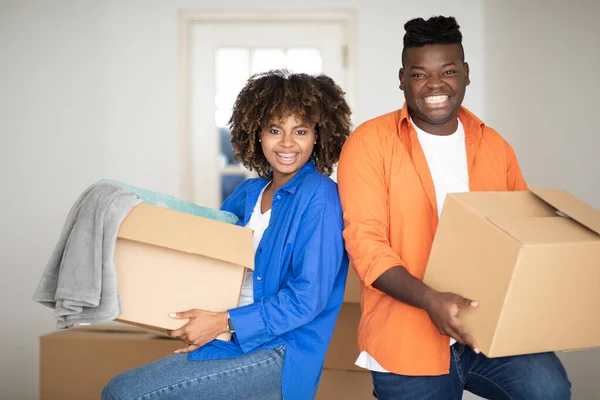 Relocation Concept. Cheerful Black Millennial Spouses Carrying Cardboard Boxes And Smiling At Camera, Happy African American Couple Packing Belongings For Moving To Their New Flat, Free Space