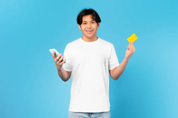 モバイルショッピング 青い背景にスタジオに立ってカメラに携帯電話の笑顔を使用して支払いを行うクレジットカードを保持する幸せなアジアのティーンエイジャーの男 Eコマース アプリケーション広告 — ストック写真