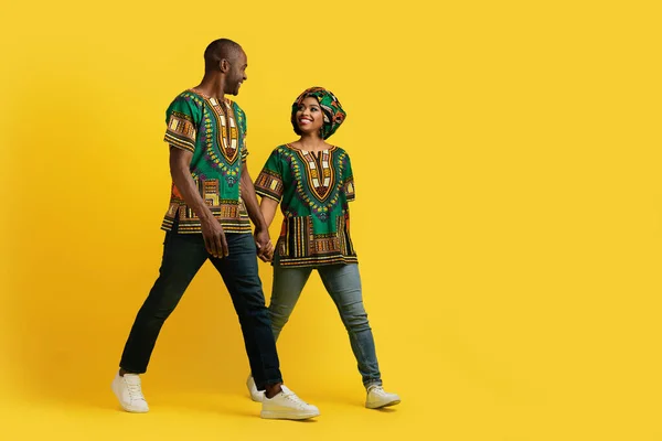 黄色のスタジオの背景に広告のためのコピースペースに向かって歩いて 手を取り合って伝統的な服を着て幸せな陽気なアフリカの男性と女性を愛し お互いに笑顔 フル長さのショット — ストック写真