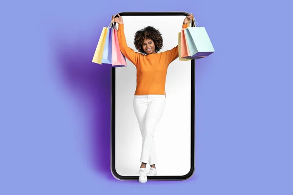 年轻而快乐的黑人女人 穿着休闲装 满头浓密的发式购物狂 站在大手机前 白色的空屏幕 微笑着 提着购物袋 紫色背景 复制空间 — 图库照片