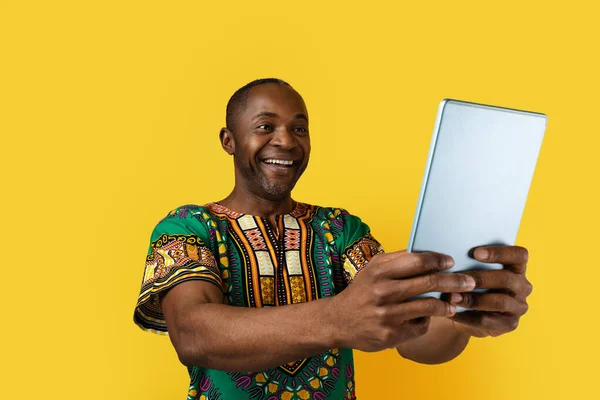 幸せな興奮ハンサムな中年黒男身に着けている伝統的なアフリカの衣装を保持します現代的なデジタルタブレットで彼の手と笑顔 黄色のスタジオの背景に隔離 — ストック写真